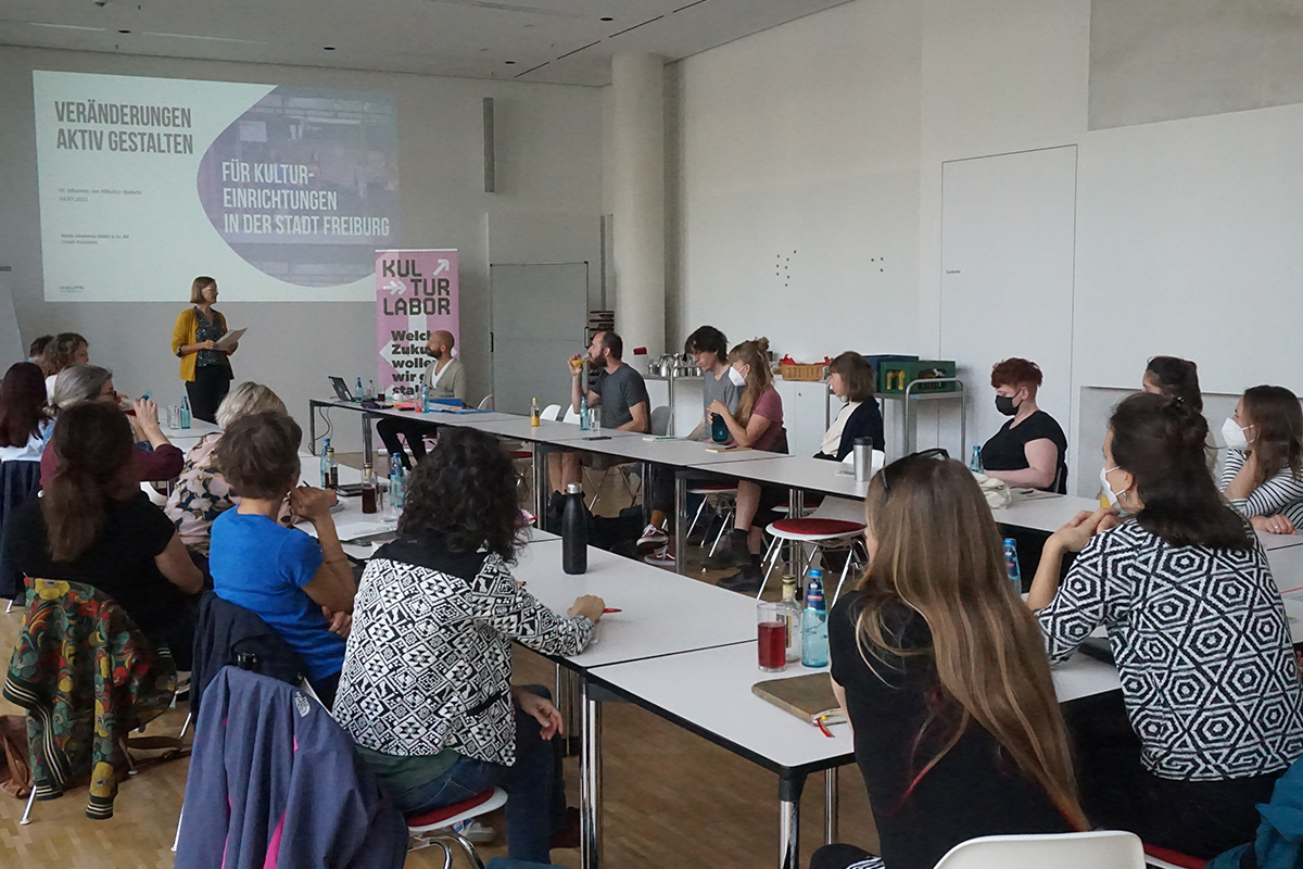 Menschen sitzen an einem u-förmigen Tisch zusammen und hören einem Vortrag zu