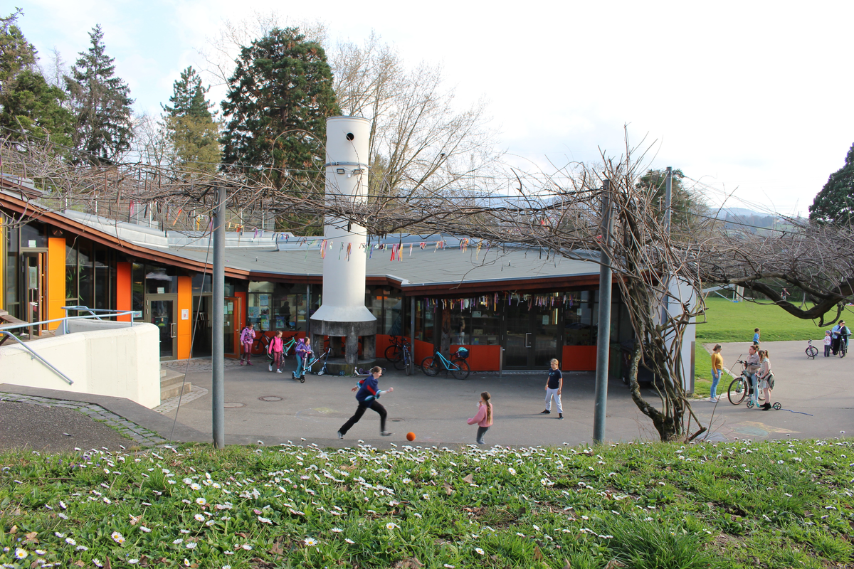 Orangenes Gebäude mit großen Fenstern und Flachdach. Spielende Kinder im Vordergrund