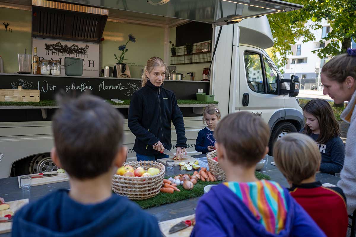 Frau schneidet Gemüse, Kinder schauen zu, im Hintergrund Foodtruck