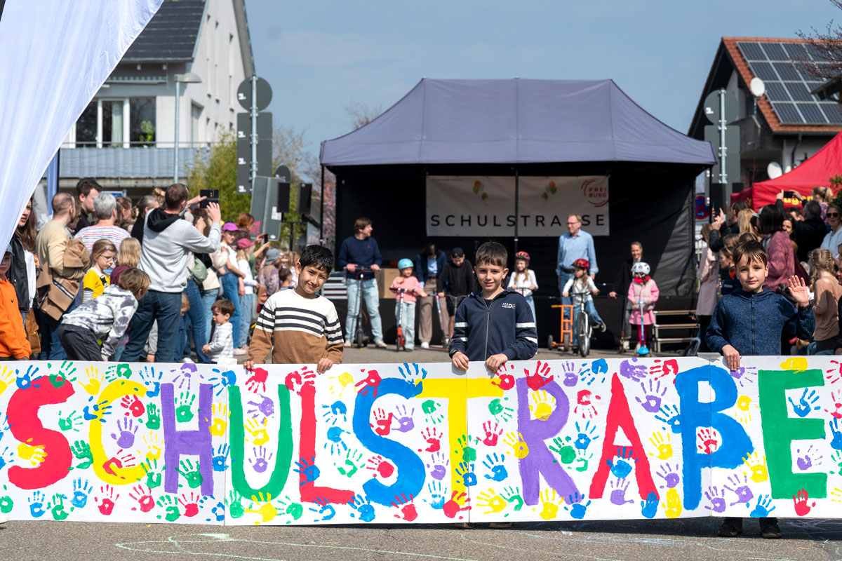 Kinder halten bei einem Straßenfest ein selbstgemaltes Banner mit der Aufschrift "Schulstraße"