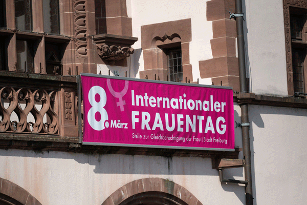 Weiße Schrift "8. März" auf Rosa Hintergrund, dahinter Hellrosa eine Zeichen für Frau