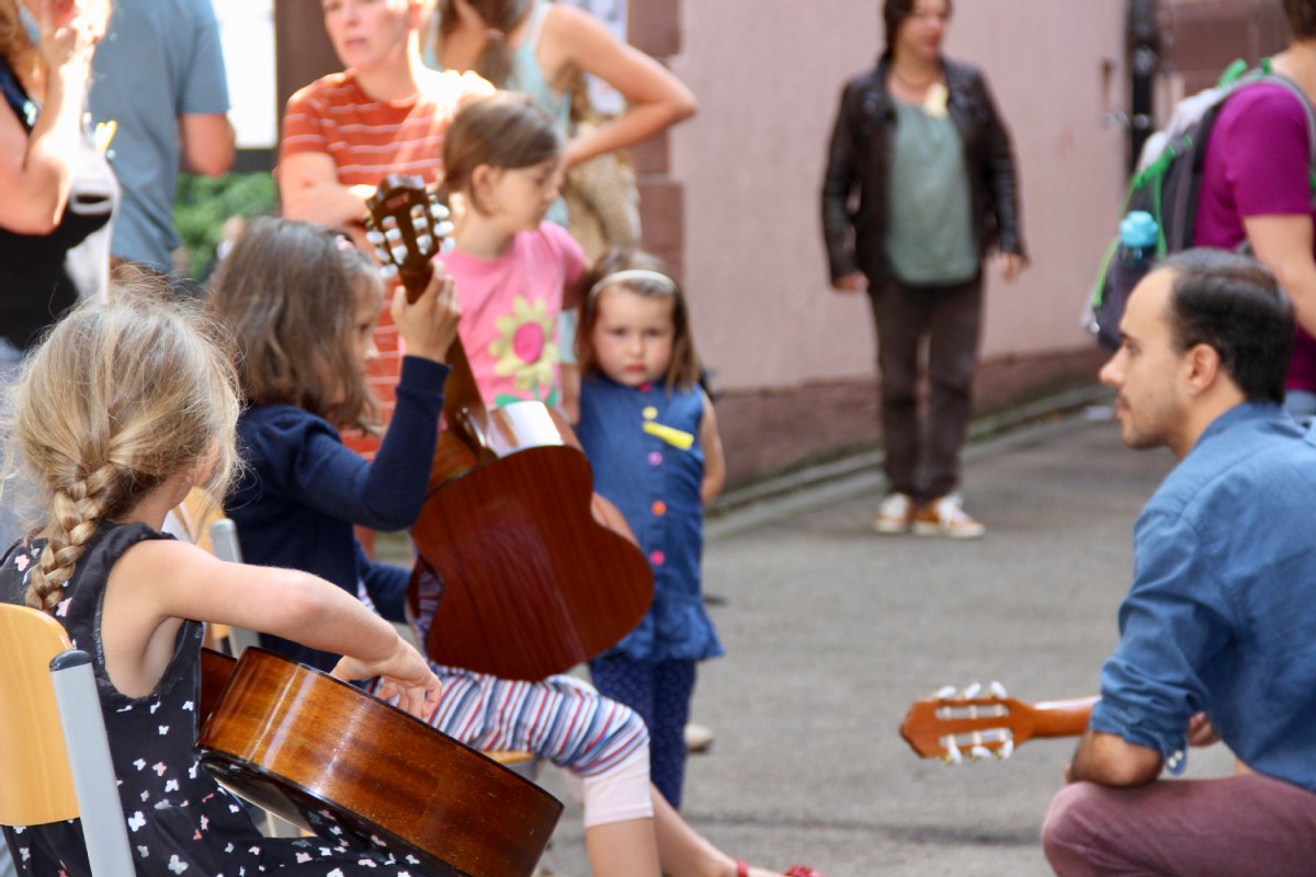 Draußen spielen Kinder und ein Erwachsener Gitarre, dahiner stehen Leute im Freien.
