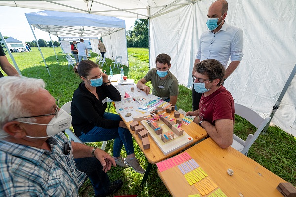 Vier Menschen mit Maske sitzen um einen Tisch mit Holzklötzchen und Aufklebern