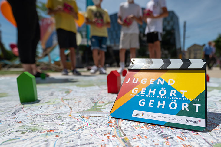 Logo der Aktion "Jugend gehört gehört", es sind zudem vier Jugendliche verschwommen sichtbar, die im Austausch mit einer Fachkraft stehen