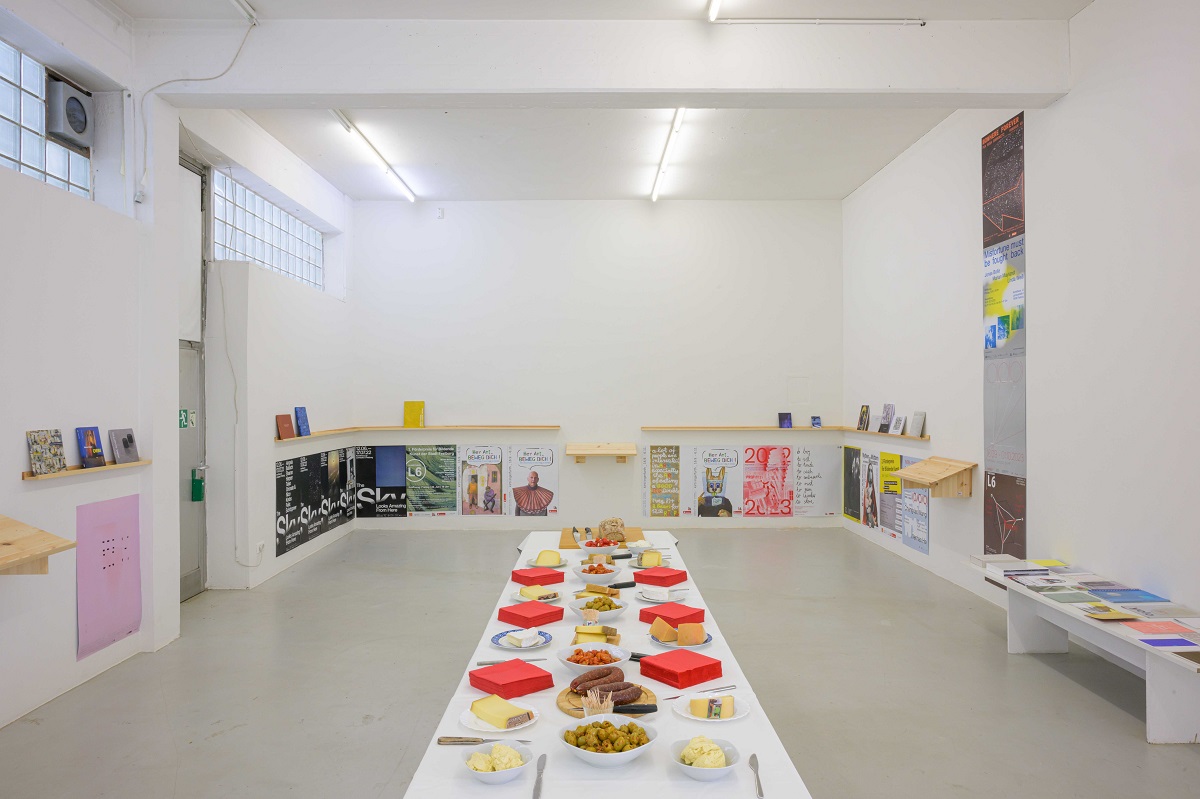 Ausstellungsansicht von "Abschied vom L6". Über Plakaten von vergangenen Ausstellungen befinden sich schamle Regale auf denen von Künstler_innen mitgebrachte Kataloge präsentiert werden. In der Mitte ist ein Tisch mit Buffet zu sehen