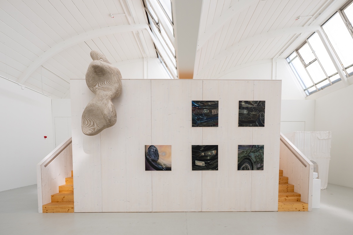 Ausstellungsansicht mit Malereien der Preisträgerin Morena Denigro (Nahansichten von Autos mit vielen Spiegelungen) links daneben eine ortspezifische große Skulptur von Luis Roether (geschwungene Form über die Empore herausragend aus Pappe)