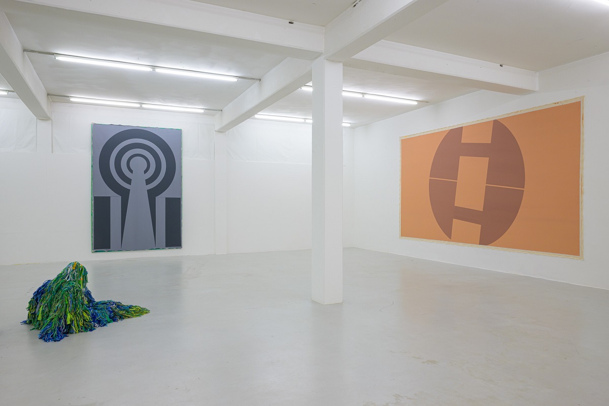 Ausstellungsansicht mit großen Malereien die an Wappen oder Symbole erinnern von Pia Rosa Dobrowitz und eine kauernde Figur  in einem blau-grünen Tarnanzug von Yongkuk Ko