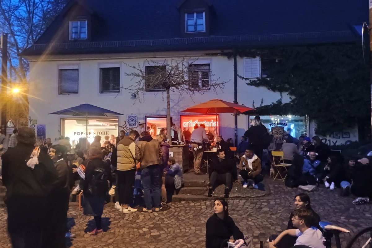 Abendstimmung - Menschen stehen an Stehtischen auf einem Platz vor einem Haus mit beleuchteten Fenstern eines Ladenlokals