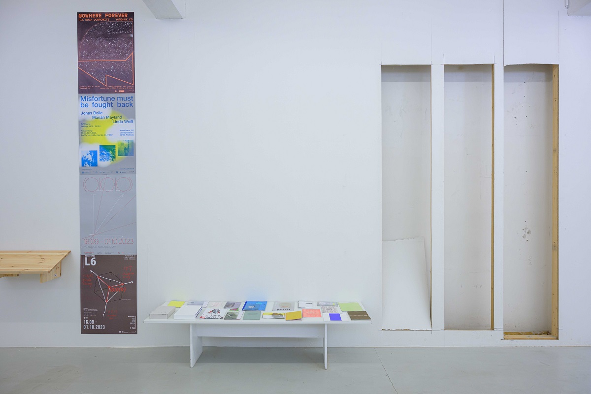 Ausstellungsansicht von "Abschied vom L6". Ein niedriger Tisch zeigt Publikationen zu Ausstellungen des Kunsthaus L6 der vergangenen 20 Jahre. Danaeneben sieht man Ausschnitte aus der Wandverkleidung aus denen die Regalkuben gebaut wurden.