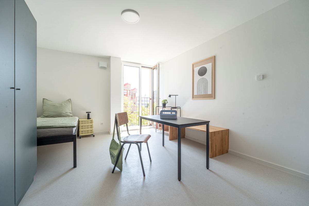 Ein Zimmer im Azubiwohnheim mit großem Fenster, Bett, Tisch, Stuhl und Sitzbank.