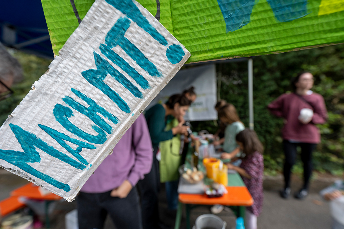 Im Vordergrund ein Pappschild mit der Aufschrift: Mach mit! Im Hintergrund sitzen Menschen an einem Tisch und verkaufen etwas.