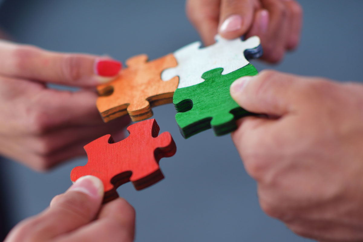 Vier Hände halten vier verschiedenfarbige Puzzleteile aneinander.