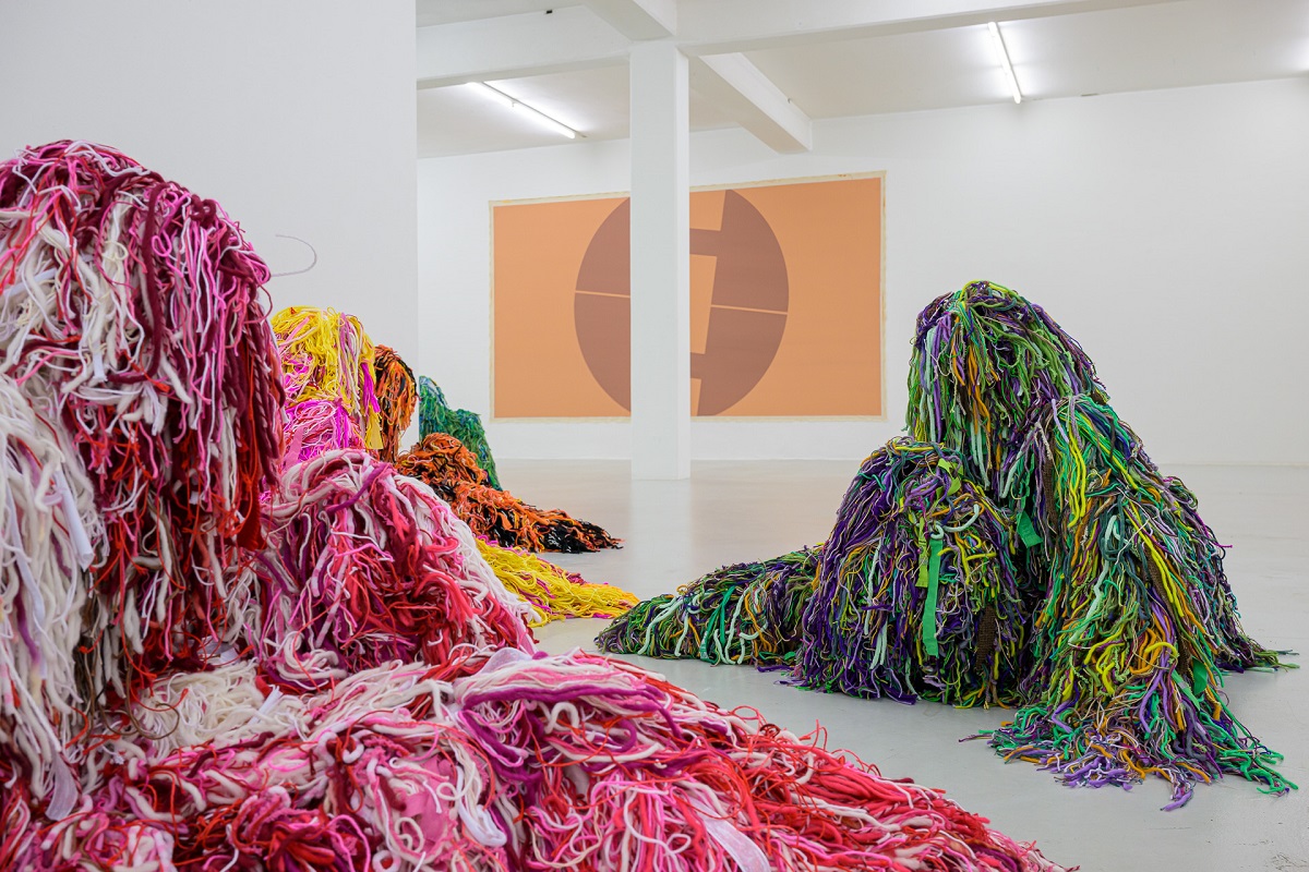 Ausstellungsansicht, Detail mit kauernden Figuren in bunten Tarnanzügen von Yongkuk Ko und im Hintergrung eine große Malerei die an ein Symbol erinnert von Pia Rosa Dobrowitz 