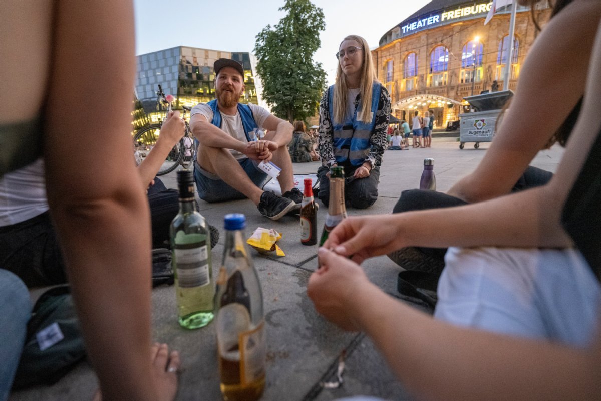 Sitzkreis, mehrere geöffnete Flaschen mit unterschiedlichen alkoholischen Getränken. Zwei Prärie-Mitarbeitende in hellblauen Warnwesten sprechen mit jungen Leuten.
