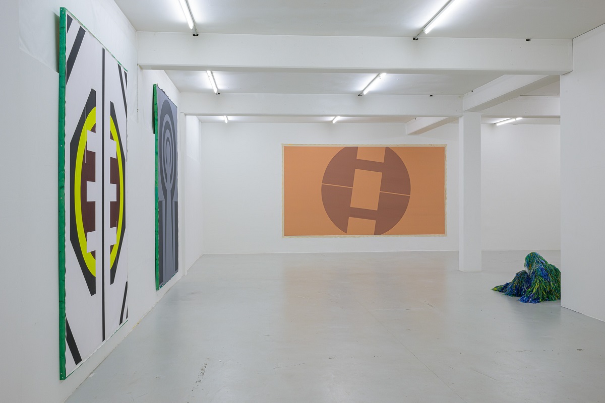 Ausstellungsansicht mit großen Malereien die an Wappen erinnern von Pia Rosa Dobrowitz und eine kauernde Figur in im Tarnanzug von Yongkuk Ko